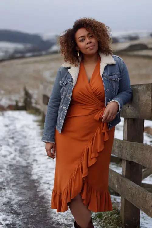 A curvy woman over 30 wears a burnt orange wrap dress with a midi hemline and ruffle detail, paired with a shearling-lined denim jacket and heeled western boots