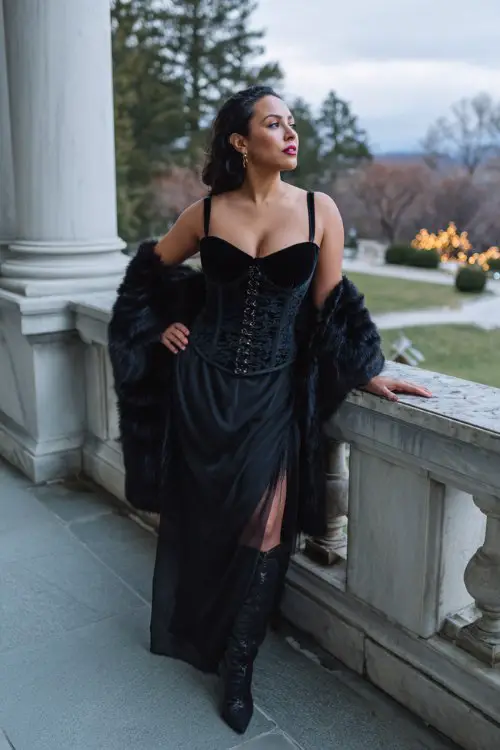 A curvy woman wears a black velvet corset-bodice gown with a square neckline and a sheer tulle overskirt, draped in a fur-trimmed satin capelet and paired with heeled boots