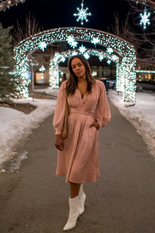 A curvy woman wears a blush pink pleated dress with long bishop sleeves and white pointed-toe ankle boots