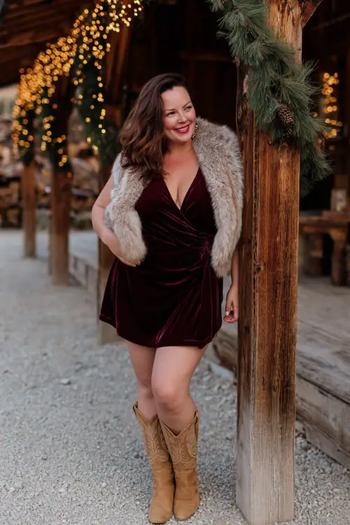A curvy woman wears a deep burgundy velvet wrap dress with tan cowboy boots and a faux fur stole