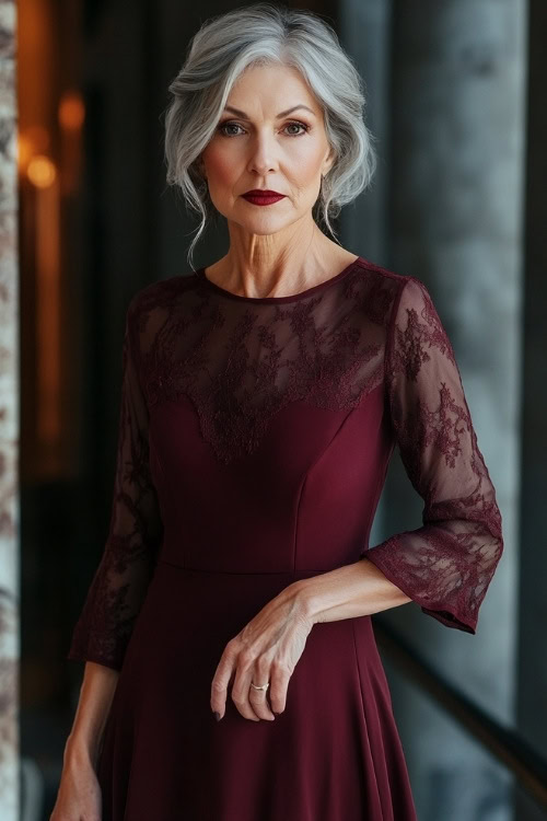 A fashionable woman over 50 in a knee-length burgundy dress with sheer sleeves, posing confidently indoors at a wedding venue (2)