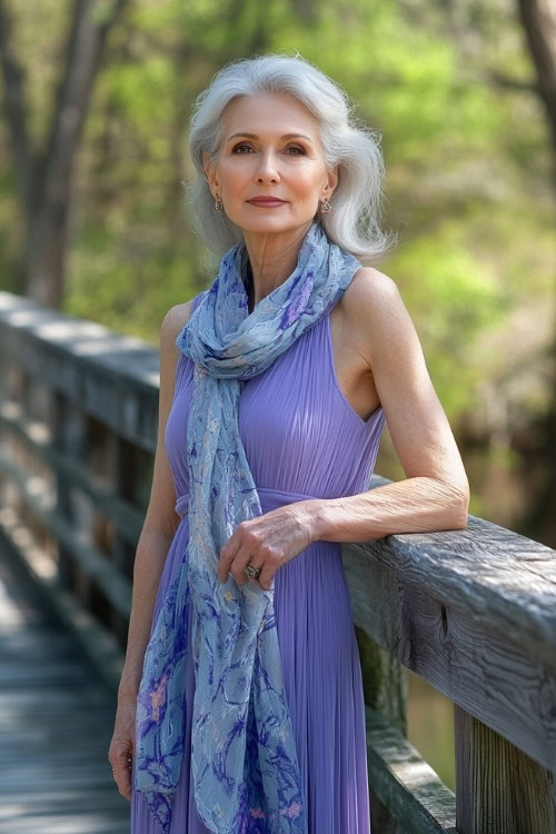 A fashionable woman over 50 wearing a periwinkle sleeveless dress with a lightweight scarf, standing by a wooden bridge in springtime