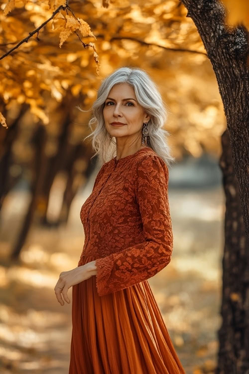 A full-body view of a woman over 50 in a rust-orange midi dress with long sleeves, standing under golden autumn trees