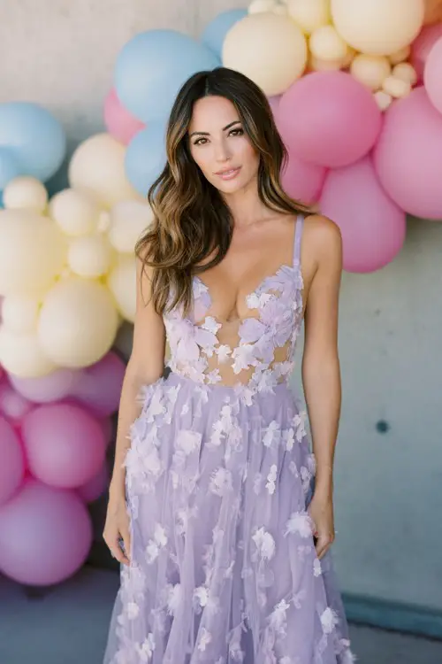 A lady in a lavender gown with delicate 3D floral appliqués scattered over a sheer overlay, standing beside a pastel balloon arch
