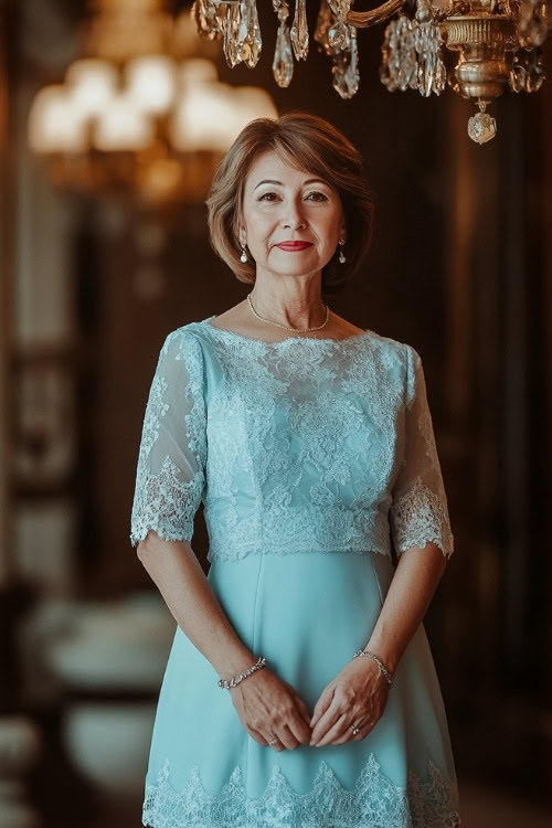 A petite woman over 50 in a light blue knee-length dress with three-quarter sleeves, standing under an ornate chandelier at a wedding