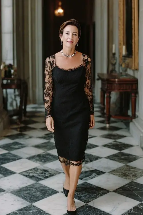 A petite woman over 50 wears a classic black midi dress with sheer lace long sleeves, styled with a crystal necklace and black heels