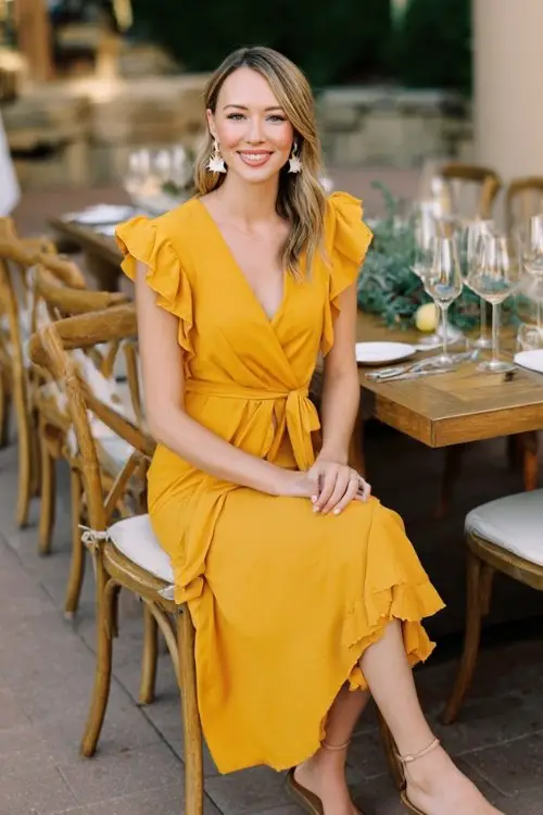 A petite woman wears a golden yellow midi wrap dress with ruffle trim, short flutter sleeves, and a waist-tie belt, paired with neutral sandals and drop earrings