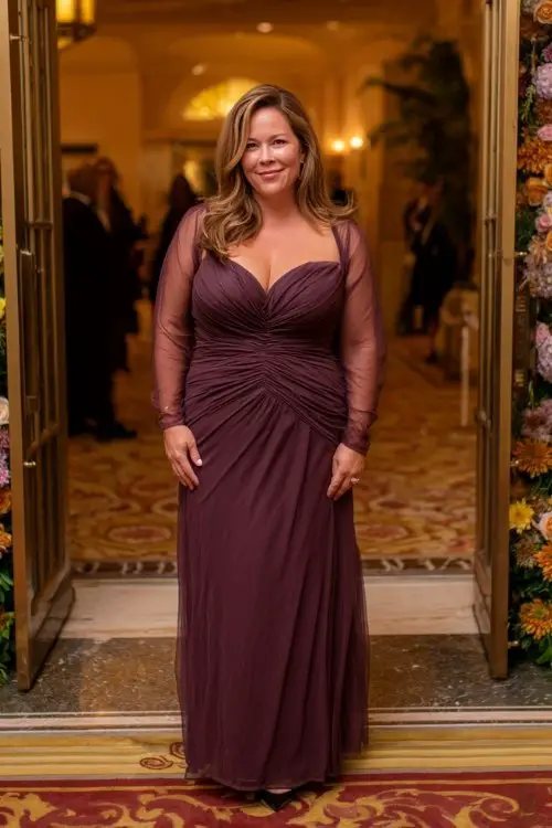 A plus-size woman over 40 wears a deep plum stretch gown with elegant ruching along the waist, sheer illusion sleeves, and a gathered neckline