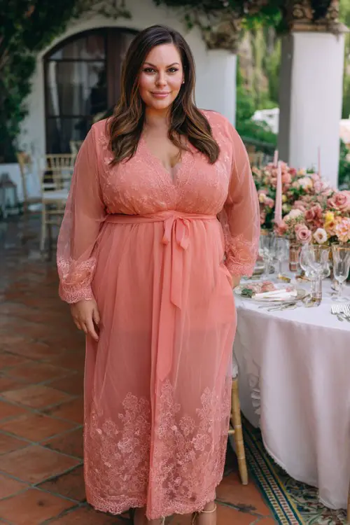 A plus-size woman over 40 wears a soft coral dress with a sheer embroidered front panel, sheer sleeves, and ribbon belt