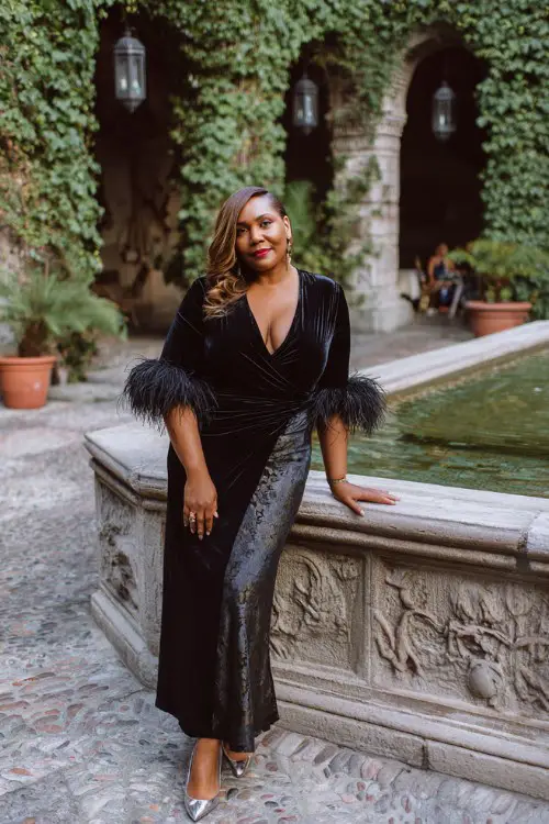 A plus-size woman over 50 wears a black velvet wrap gown with a satin panel skirt and dramatic feather-trim cuffs, paired with metallic heels