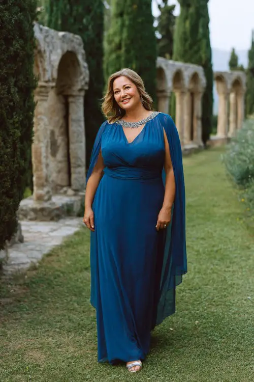 A plus-size woman over 50 wears a floor-length sapphire blue gown with a flowing chiffon cape attached at the shoulders, subtle crystal beading on the neckline, and silver heels