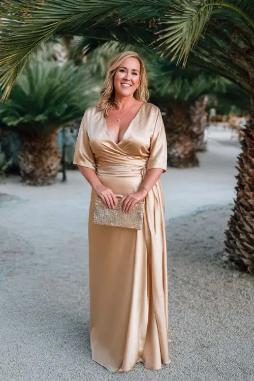 A plus-size woman over 50 wears a golden satin gown with a soft cowl neckline, flowy skirt, and beaded clutch