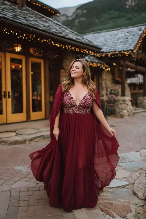A plus-size woman wears a burgundy A-line gown with an embroidered bodice and chiffon cape flowing in the breeze