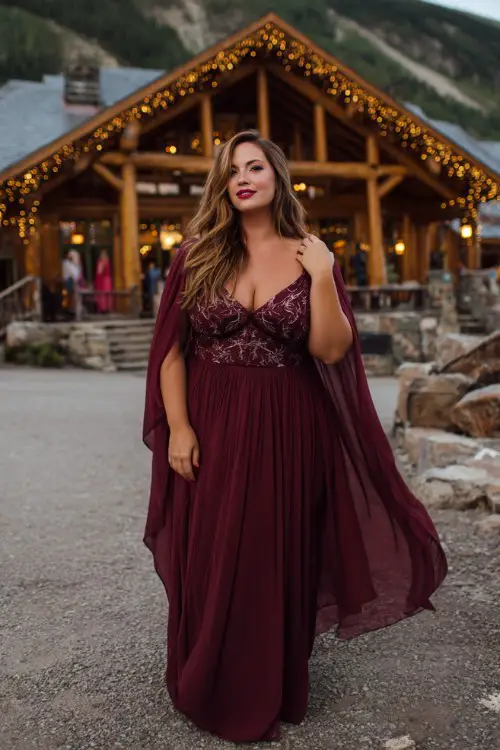 A plus-size woman wears a burgundy A-line gown with an embroidered bodice and chiffon cape flowing in the breeze