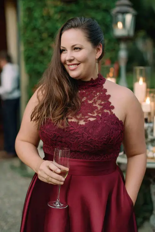 A plus-size woman wears a deep red dress with a high neckline, lace bodice, and satin flare skirt