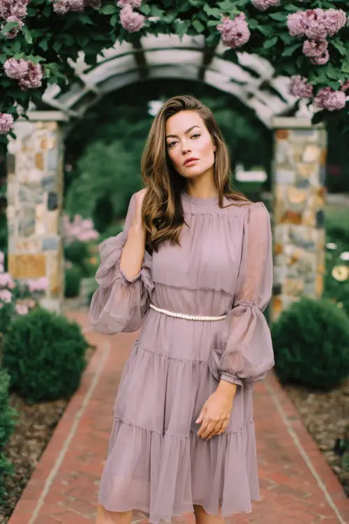 A plus-size woman wears a dusty lavender midi dress with tiered layers, sheer poet sleeves, and a pearl belt detail