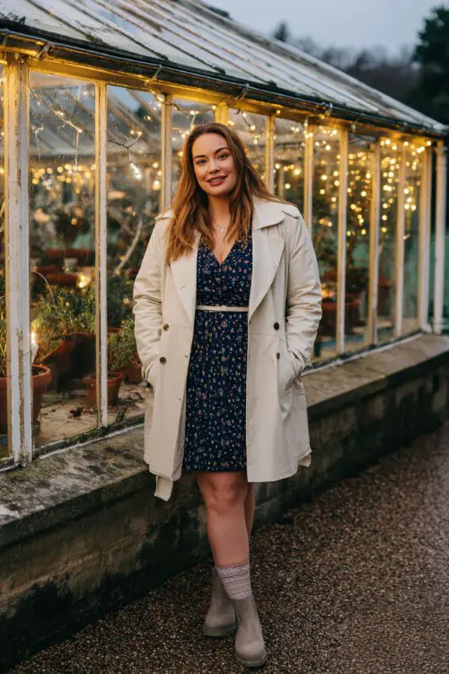 A plus-size woman wears a navy A-line dress with metallic embroidery and matching sock boots, layered under a cream belted coat