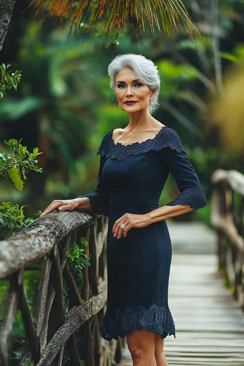 A stylish woman over 50 in a navy cocktail dress with a scalloped hemline and elbow-length sleeves, posing by a wooden bridge in a garden wedding