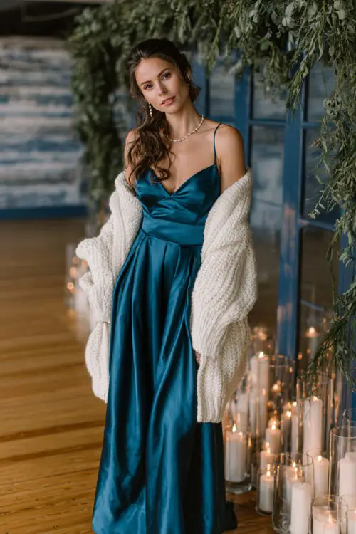 A woman in a dark teal satin gown with an oversized white cardigan and pearl accessories