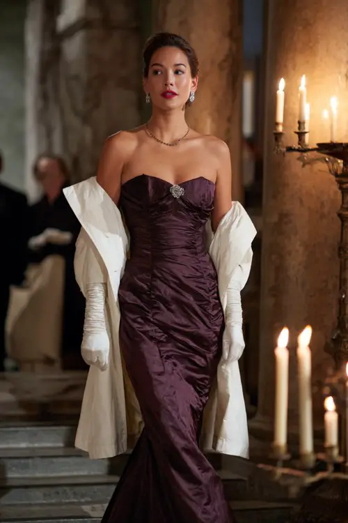 A woman over 30 wears a dark plum mermaid gown with structured pleats and opera gloves