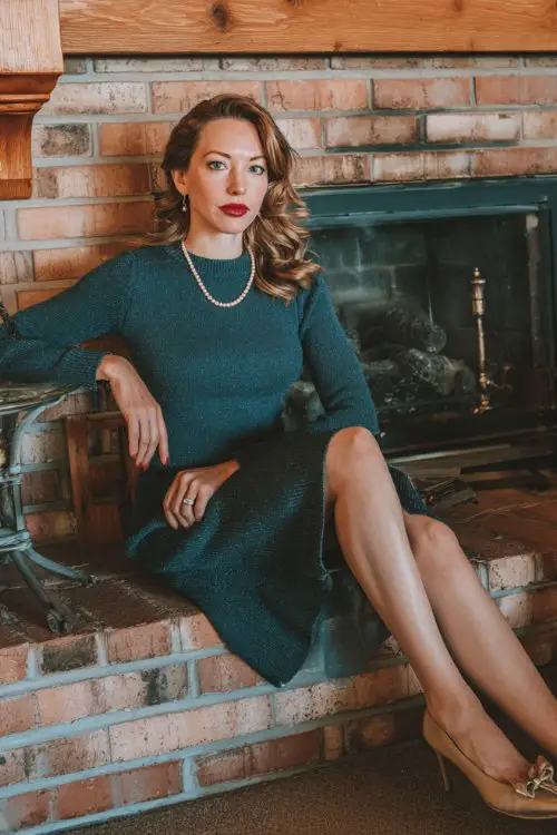 A woman over 30 wears a dark teal sweater dress with a slightly flared skirt and subtle shimmer, paired with Mary Jane shoes and pearl accessories