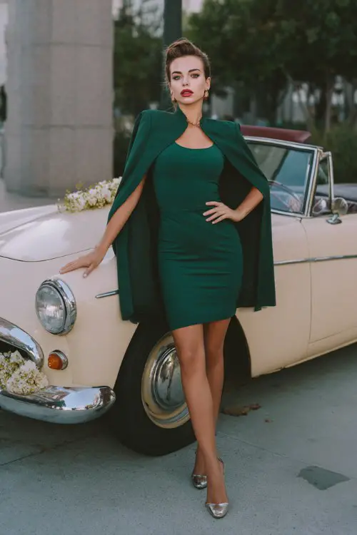 A woman over 30 wears a fitted forest green mini dress with a structured cape detail and metallic heels