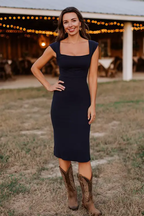 A woman over 30 wears a fitted navy midi dress with a square neckline and brown distressed cowboy boots