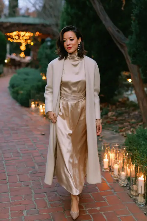 A woman over 30 wears a high-neck champagne gown with a satin skirt and subtle shimmer, topped with a long cream wool wrap coat and pearl accessories