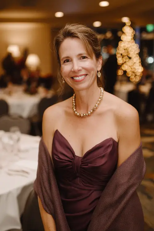 A woman over 35 wears a burgundy high-neck dress with lace sleeves and tan heeled boots, a plaid wool wrap draped around her shoulders as she walks through a candle-lit garden