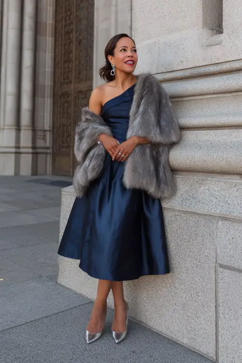 A woman over 40 wears a navy blue satin A-line dress with a dramatic one-shoulder cape sleeve, paired with silver heels and a long faux fur stole wrapped around her arms