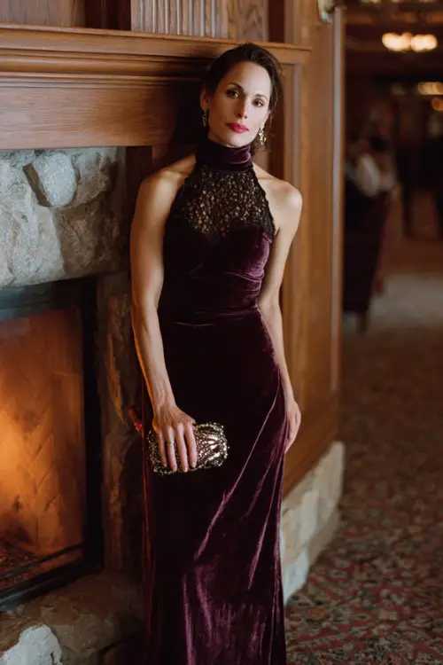 A woman over 40 wears a plum-colored high-neck velvet gown with lace insets and embellished clutch