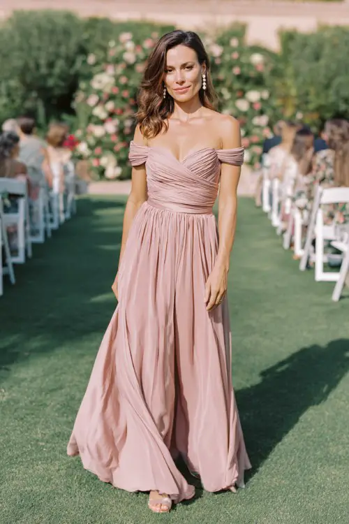 A woman over 40 wears a soft blush pink off-the-shoulder pleated gown with a satin waist tie and pearl earrings