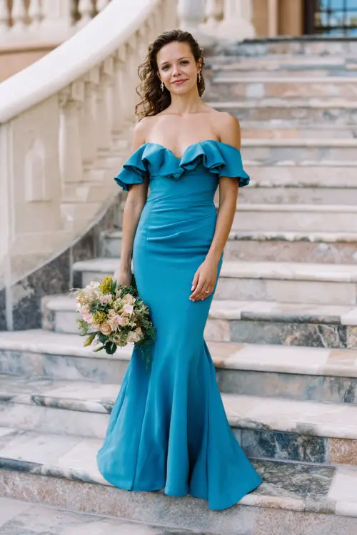 A woman over 40 wears a teal off-shoulder mermaid gown with ruffled detail and a small train