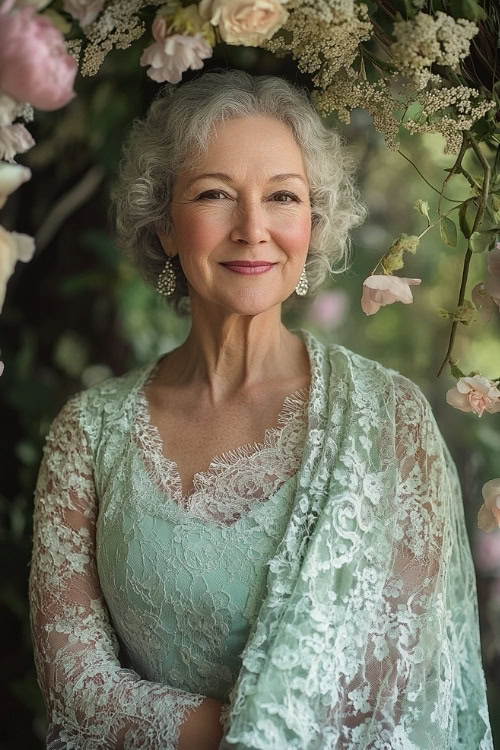 A woman over 50 in a mint green lace dress with a matching shawl, smiling under a wedding arch adorned with spring flowers