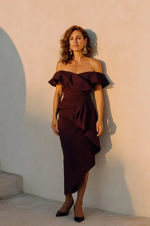 A woman over 50 wears a burgundy off-shoulder cocktail dress with asymmetrical ruffles and gold earrings