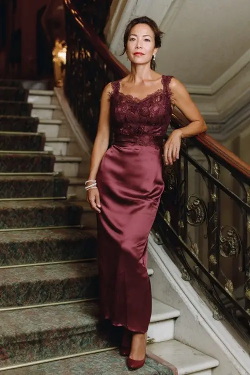 A woman over 50 wears a burgundy satin gown with a lace bodice overlay and scalloped neckline, paired with satin heels and a pearl bracelet