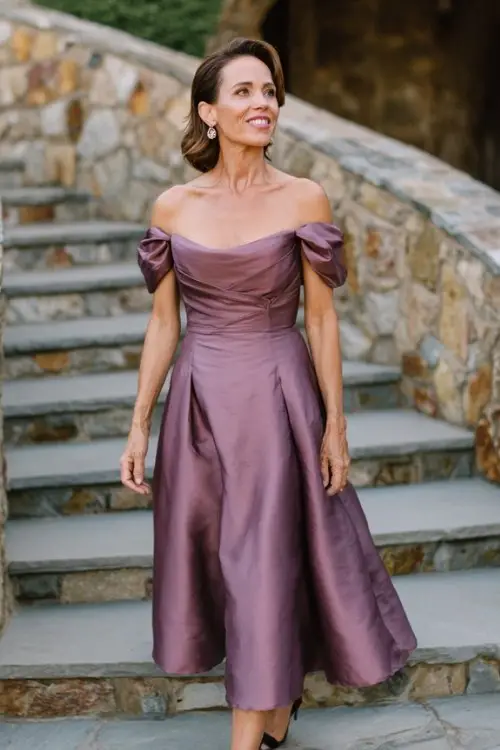 A woman over 50 wears a mauve off-the-shoulder formal dress with a structured bodice and soft flare skirt, styled with vintage drop earrings and block heels