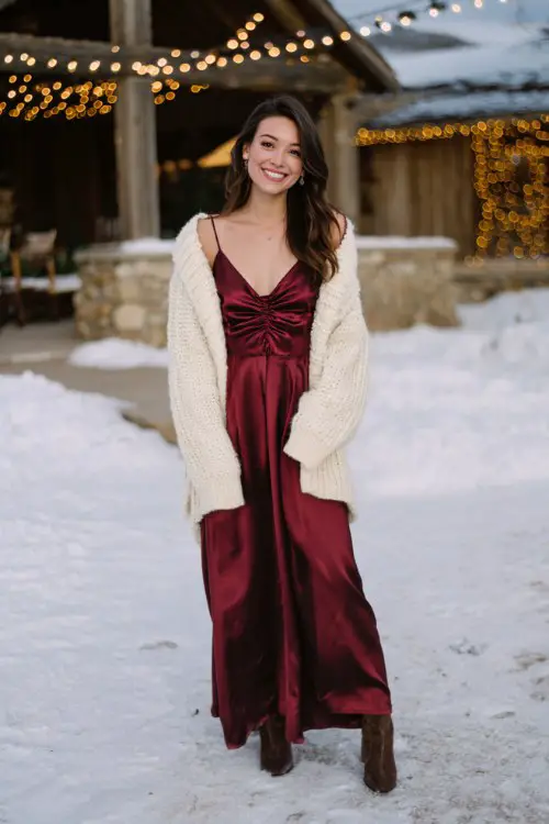 A woman wearing a long burgundy satin dress with a cream knit cardigan and heeled boots