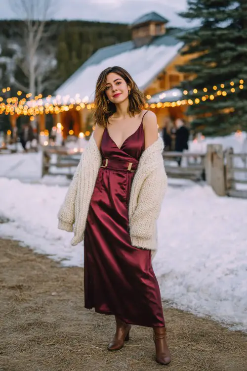 A woman wearing a long burgundy satin dress with a cream knit cardigan and heeled boots