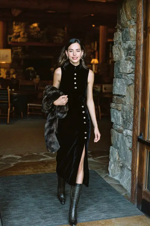 A woman wears a black velvet mock-neck midi dress with silver buttons down the back, paired with knee-high snakeskin boots and a faux fur shawl