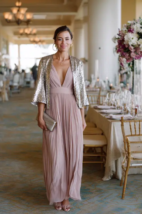 A woman wears a blush pink pleated gown with flutter sleeves and a metallic clutch, styled with a silver sequin jacket and silver heels