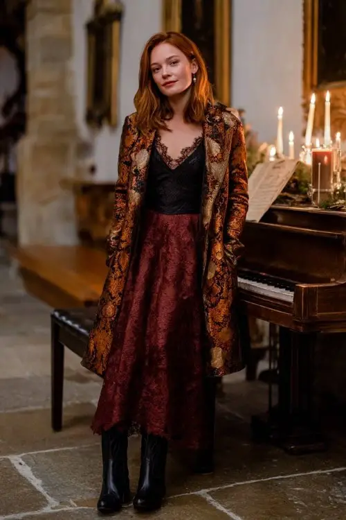 A woman wears a burgundy lace gown with sheer sleeves, a patterned jacquard coat in gold tones, and black cowboy boots