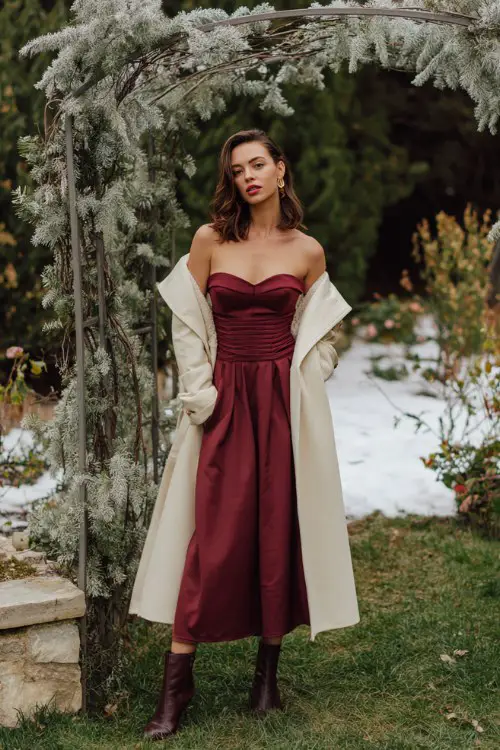 A woman wears a burgundy off-the-shoulder gown with a structured bodice, styled with heeled boots and a cream tailored coat