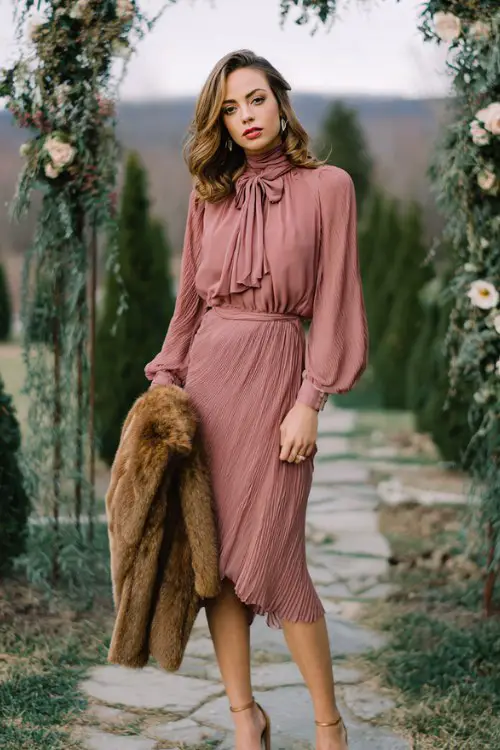 A woman wears a dusty rose long-sleeve tea-length dress with pleated chiffon and a high neckline, styled with block heels and a fuzzy shawl