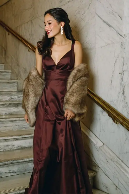 A woman wears a floor-length burgundy gown with a fitted bodice, pearl earrings, and a fur stole