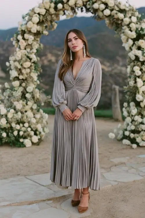 A woman wears a gray pleated dress with long sleeves, silver earrings, and suede heels