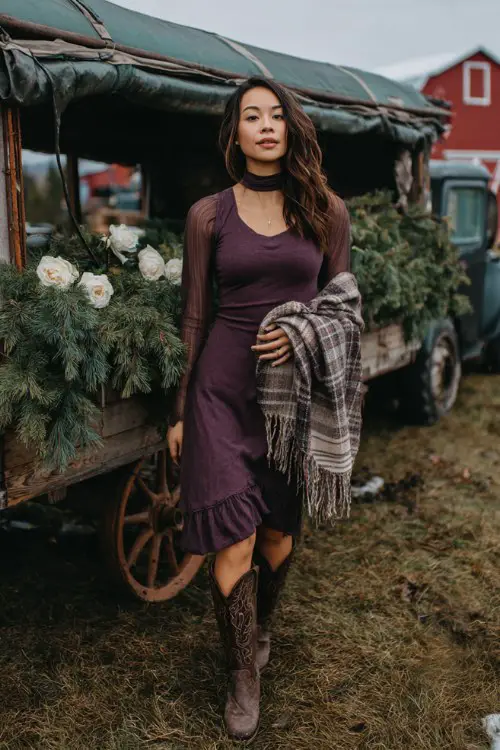 A woman wears a high-neck fitted dress in plum with sheer sleeves, a plaid blanket shawl, and distressed cowboy boots