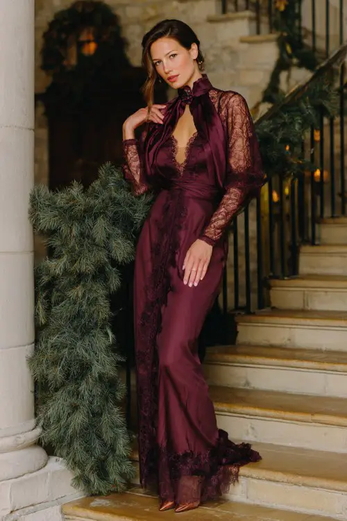 A woman wears a high-neck long-sleeve gown in deep wine with sheer lace detailing, styled with a velvet embroidered shawl and pointed heels