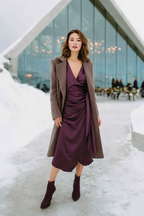 A woman wears a long-sleeve deep plum satin midi dress with a draped bodice, paired with a belted wool coat and heeled ankle boots