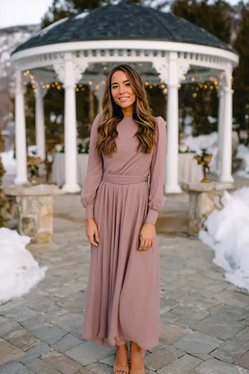 A woman wears a long-sleeved chiffon maxi dress in dusty rose, soft waves in her hair, and neutral block heels